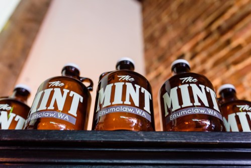 Brown glass bottles labeled "The Mint, Enumclaw, WA" arranged on a shelf against a brick wall.