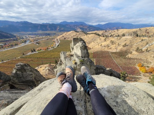 Two pairs of feet in hiking boots resting on a rocky ledge, overlooking a valley with vineyards and mountains in the background.