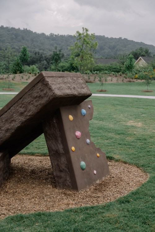 A large climbing wall with colorful holds, set in a grassy park with trees and a cloudy sky in the background.