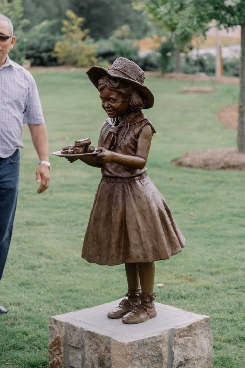 A bronze statue of a young girl in a dress and hat, holding a plate, stands on a stone base in a grassy area.