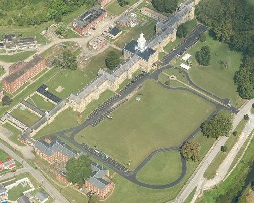 Aerial view of a large, historic building complex with manicured lawns and parking areas, surrounded by trees.