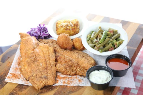 A plate of fried fish fillets with hushpuppies, green beans, cornbread, and dipping sauces on a wooden table.