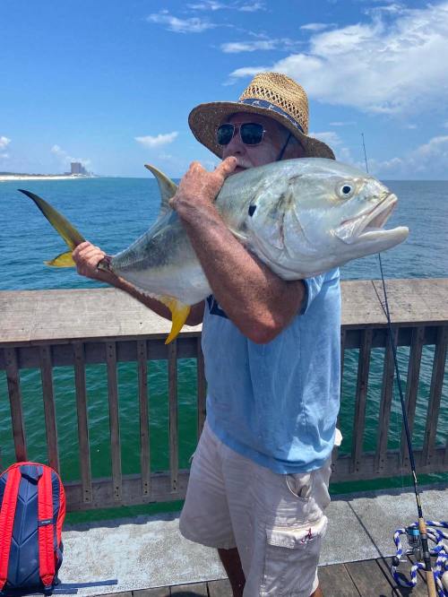 A person in a straw hat holds a large fish while standing on a pier by the water.