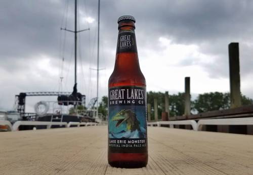A bottle of Great Lakes Brewing Co.'s Lake Erie Monster IPA on a wooden dock with boats in the background under cloudy skies.