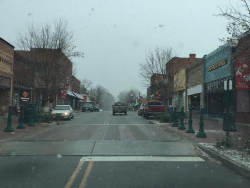 Snow falls on a quiet downtown street lined with shops and parked cars, creating a serene winter scene.
