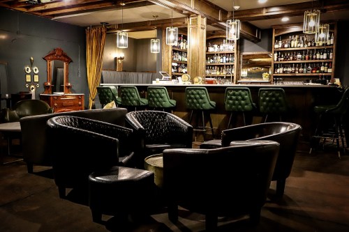 Cozy bar interior featuring leather chairs, a wooden counter, and shelves stocked with various bottles. Warm lighting.