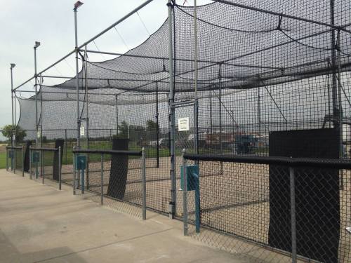 A view of a baseball batting cage with nets and a concrete pathway, set in an outdoor sports facility.
