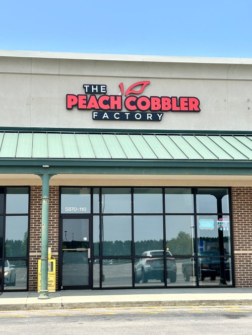 Sign for "The Peach Cobbler Factory" on a storefront with a green awning and large glass windows.