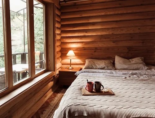 Cozy cabin interior with a wooden bed, two mugs on a tray, and large windows overlooking nature.