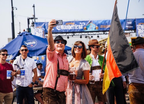 A group of people in traditional attire take a selfie at an Oktoberfest celebration, holding drinks and smiling.
