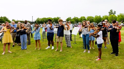 A group of young musicians playing violins outdoors, with a diverse audience in the background.