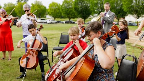 A group of children playing string instruments outdoors, with an adult musician in the background.