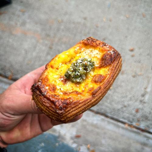 A hand holding a golden pastry topped with green sauce and sprinkled with cheese, set against a concrete background.