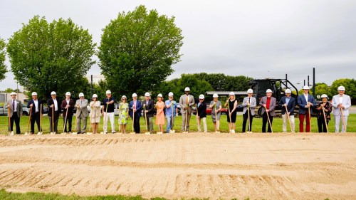 A group of people in hard hats and suits pose for a groundbreaking ceremony on a sandy site with trees in the background.
