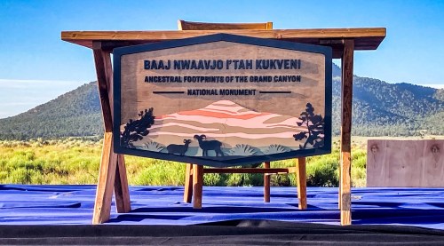 A wooden sign reading "Baaj Nwaavjo I'tah Kukveni" in front of a scenic landscape with mountains and blue sky.