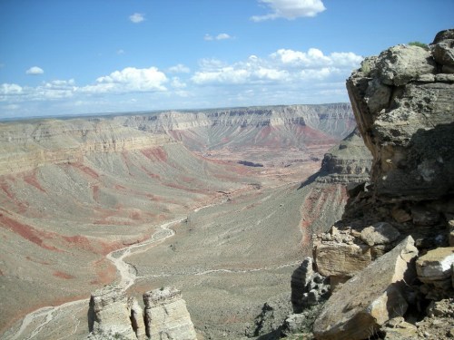 A vast canyon landscape with layered rock formations, a winding river, and a blue sky dotted with clouds.