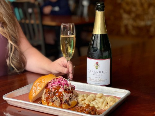 A plate with a pulled pork sandwich, macaroni, and a glass of sparkling wine beside a bottle of Greenhill Vineyards.