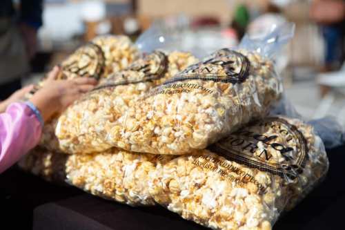 A stack of large bags filled with popcorn on a table, with a hand reaching towards the top bag.