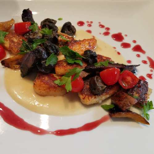 A gourmet dish featuring seared gnocchi, cherry tomatoes, mushrooms, and a drizzle of sauce on a white plate.
