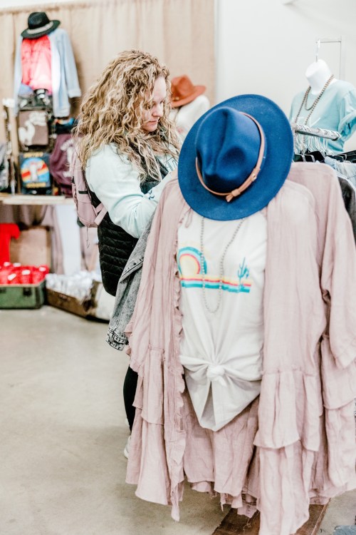 A woman browses clothing on a rack in a boutique, with a blue hat and a pink garment in focus.