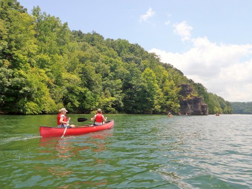 Two people in a red canoe paddle on a calm lake surrounded by lush green trees and rocky cliffs.