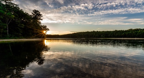 Sunset over a calm lake, with reflections on the water and trees lining the shore under a partly cloudy sky.