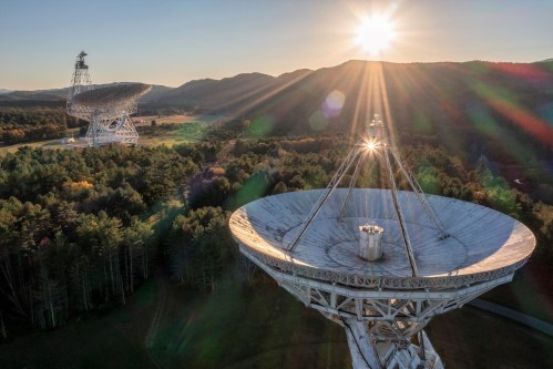 Aerial view of large satellite dishes in a forested area, with the sun setting behind mountains in the background.