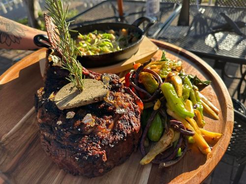 A large, grilled steak garnished with rosemary, served with colorful sautéed vegetables on a wooden platter.