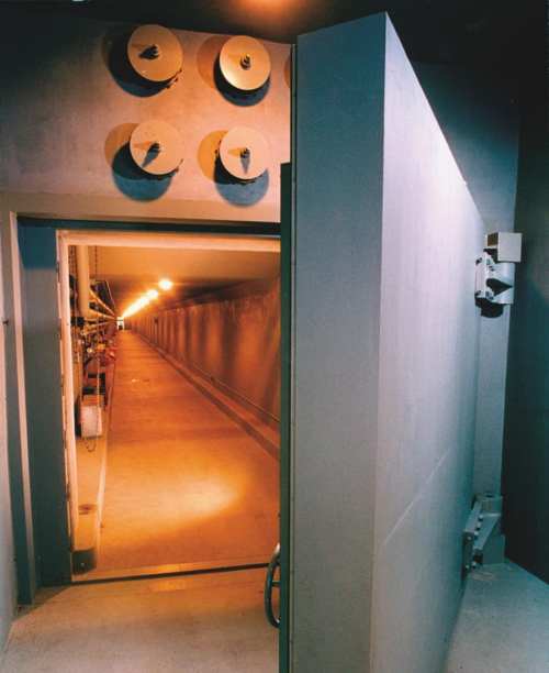 A dimly lit corridor with a large, heavy door partially open, revealing a long, empty passageway.