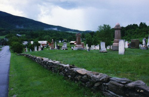 A cemetery with various gravestones, surrounded by green grass and mountains under a cloudy sky.
