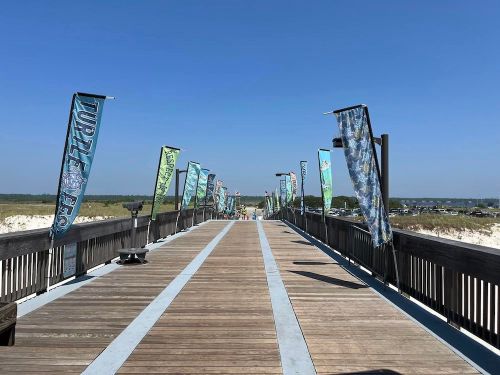 A wooden boardwalk lined with colorful flags, leading towards a sunny beach and clear blue sky.
