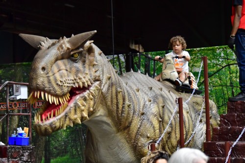 A child sits on a large dinosaur statue, smiling, with a forest backdrop and people in the background.