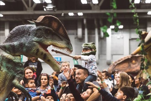 A crowd of excited children and adults interact with life-sized dinosaur models at a lively event.