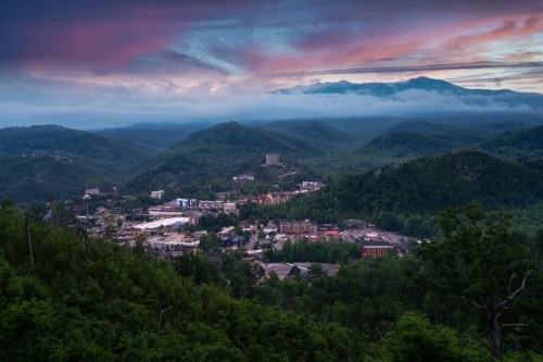 A scenic view of a mountain town surrounded by lush greenery and misty mountains under a colorful sunset sky.