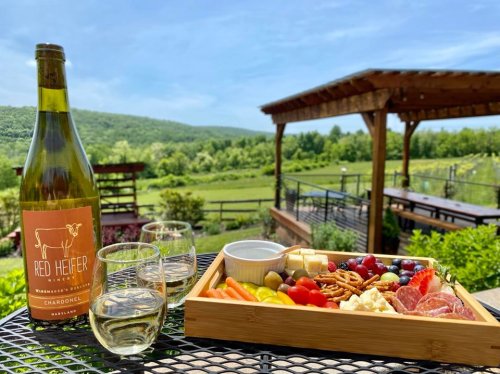 A wooden platter with snacks and wine glasses, set against a scenic outdoor backdrop of greenery and blue skies.