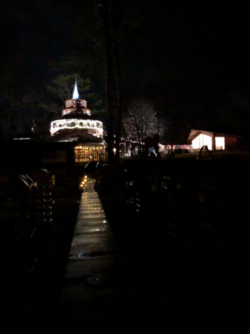 A dimly lit pathway leads to a brightly illuminated building at night, surrounded by trees.