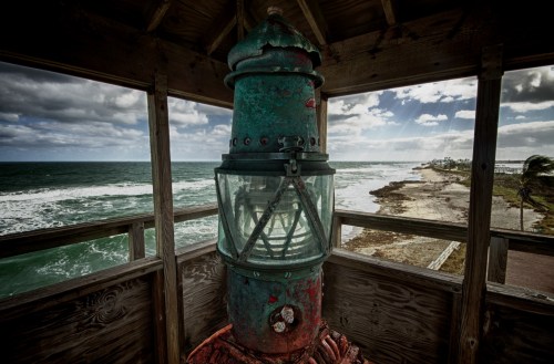 A weathered lighthouse lantern overlooking a stormy sea and coastline under a cloudy sky.
