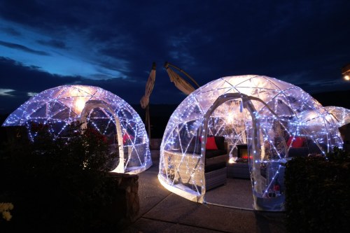 Two illuminated igloo-shaped structures with fairy lights, surrounded by plants, under a dramatic evening sky.