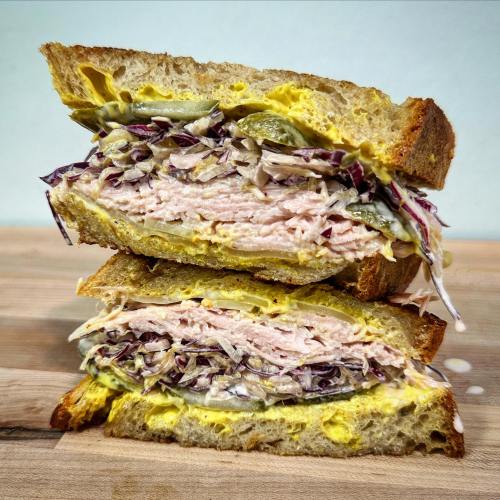 A stacked sandwich with layers of turkey, coleslaw, pickles, and mustard on whole grain bread.