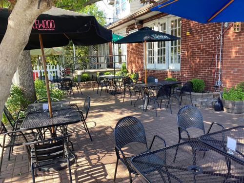 Outdoor dining area with black metal tables and chairs, shaded by umbrellas, surrounded by greenery and brick walls.
