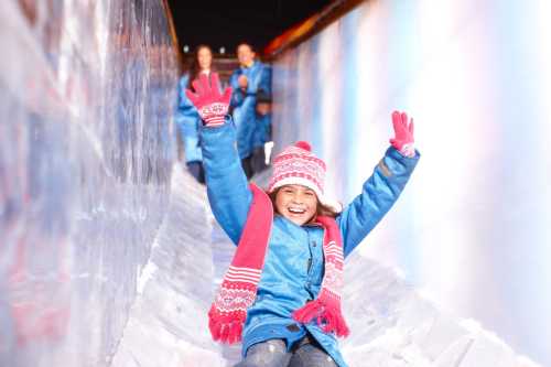 A joyful girl in winter attire slides down an ice slide, raising her hands in excitement.