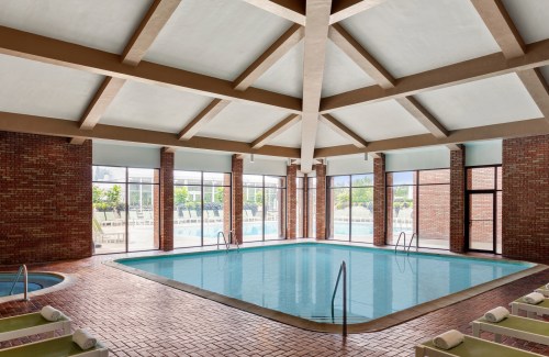 Indoor swimming pool with large windows, brick walls, and a high ceiling, surrounded by lounge chairs.