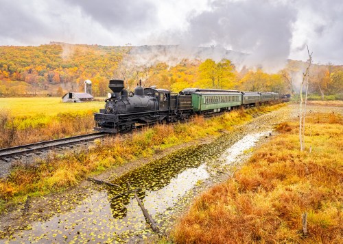 A vintage steam train travels through a colorful autumn landscape, reflecting in a nearby pond.