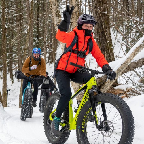 Two cyclists ride through a snowy forest trail, one waving and wearing bright winter gear, while the other follows behind.
