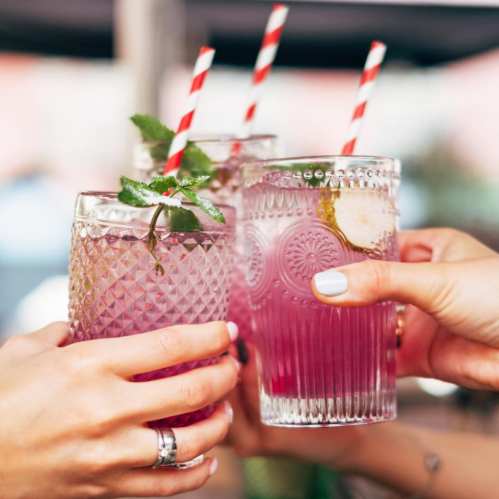 Three hands holding colorful drinks with mint and striped straws, clinking glasses in a celebratory toast.