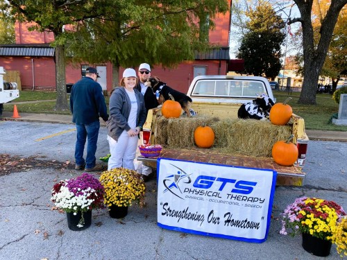 A woman and a man stand by a pickup truck decorated with pumpkins and flowers, promoting GTS Physical Therapy.
