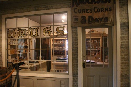 A vintage drugstore window displaying the word "DRUGS" and a sign advertising a corn cure.