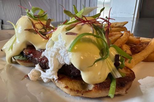 Two poached eggs on a toasted bun with greens and hollandaise sauce, served with a side of fries.