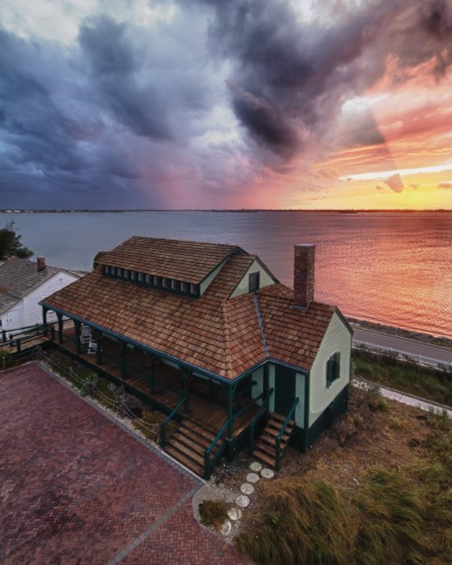 A charming house by the water at sunset, with dramatic clouds and colorful sky reflecting on the calm surface.