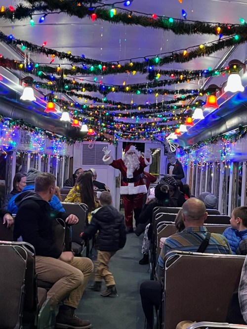 A festive train interior decorated with colorful lights, featuring Santa Claus entertaining passengers.